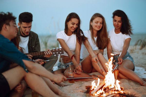 Group of young friends sitting on beach and fry sausages. One man is playing guitar. Camping time.
